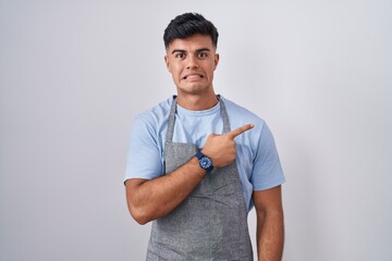 Sticker - Hispanic young man wearing apron over white background pointing aside worried and nervous with forefinger, concerned and surprised expression