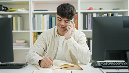 Wall Mural - Young hispanic man student talking on smartphone writing notes at library university