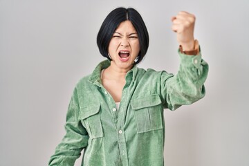 Wall Mural - Young asian woman standing over white background angry and mad raising fist frustrated and furious while shouting with anger. rage and aggressive concept.