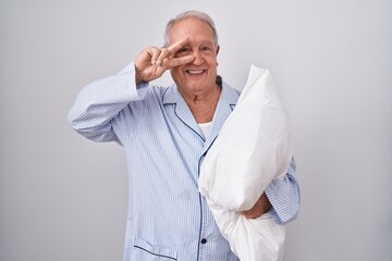 Poster - Senior man with grey hair wearing pijama hugging pillow doing peace symbol with fingers over face, smiling cheerful showing victory