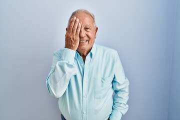 Senior man with grey hair standing over blue background covering one eye with hand, confident smile on face and surprise emotion.