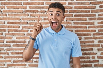 Canvas Print - Brazilian young man standing over brick wall pointing finger up with successful idea. exited and happy. number one.