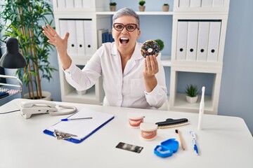 Poster - Middle age dentist woman holding chocolate doughnut smiling cheerful pointing with hand and finger up to the side