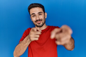 Wall Mural - Young hispanic man with beard wearing red t shirt over blue background pointing fingers to camera with happy and funny face. good energy and vibes.