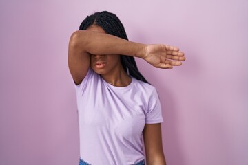 Poster - African american woman with braids standing over pink background covering eyes with arm, looking serious and sad. sightless, hiding and rejection concept