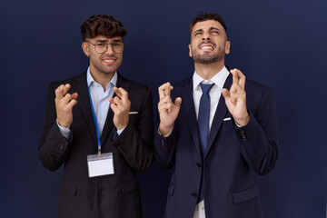 Wall Mural - Two hispanic business men wearing business clothes gesturing finger crossed smiling with hope and eyes closed. luck and superstitious concept.