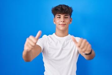 Wall Mural - Hispanic teenager standing over blue background approving doing positive gesture with hand, thumbs up smiling and happy for success. winner gesture.