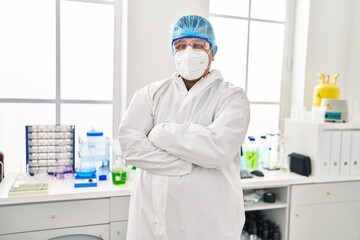 Wall Mural - Young latin man scientist wearing covid protection uniform standing with arms crossed gesture at laboratory