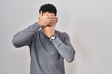 Canvas Print - Hispanic man with beard standing over white background covering eyes and mouth with hands, surprised and shocked. hiding emotion