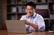 © YURII MASLAK - Asian man holding online webinar using laptop webcam, sitting in a home office. Man having online meeting and communicating remotely, looking at computer screen