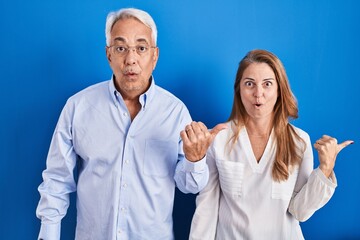 Wall Mural - Middle age hispanic couple standing over blue background surprised pointing with hand finger to the side, open mouth amazed expression.