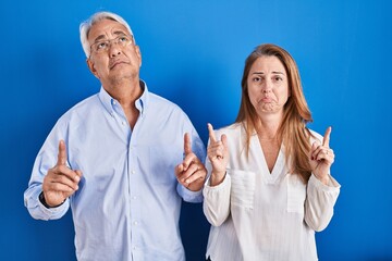 Wall Mural - Middle age hispanic couple standing over blue background pointing up looking sad and upset, indicating direction with fingers, unhappy and depressed.