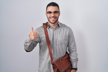 Wall Mural - Young hispanic man wearing suitcase doing happy thumbs up gesture with hand. approving expression looking at the camera showing success.