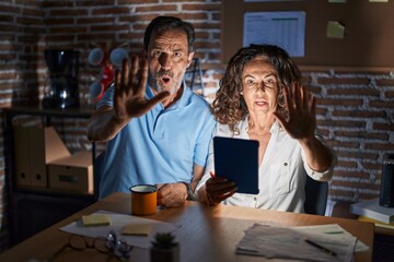 Poster - Middle age hispanic couple using touchpad sitting on the table at night doing stop gesture with hands palms, angry and frustration expression