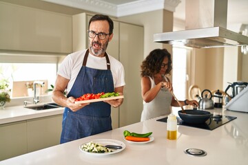 Sticker - Middle age couple cooking mediterranean food at home in shock face, looking skeptical and sarcastic, surprised with open mouth