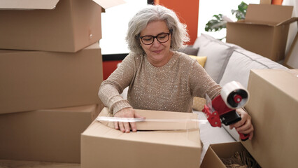 Poster - Middle age woman with grey hair smiling confident packing cardboard box at new home