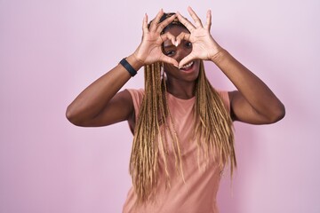 Poster - African american woman with braided hair standing over pink background doing heart shape with hand and fingers smiling looking through sign