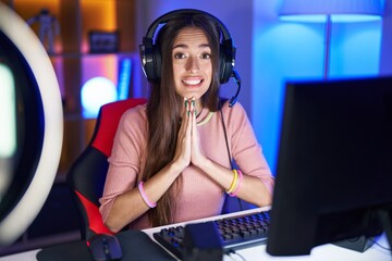 Poster - Young hispanic woman playing video games praying with hands together asking for forgiveness smiling confident.