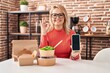 © Krakenimages.com - Blonde woman eating take away food showing smartphone screen doing ok sign with fingers, smiling friendly gesturing excellent symbol
