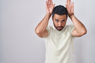 Wall Mural - Hispanic man with beard standing over isolated background doing bunny ears gesture with hands palms looking cynical and skeptical. easter rabbit concept.