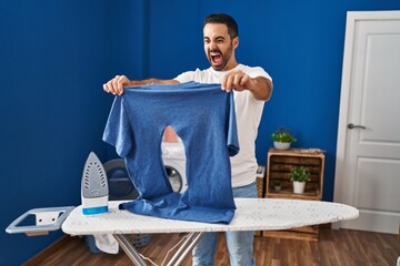 Sticker - Young hispanic man with beard ironing holding burned iron shirt at laundry room angry and mad screaming frustrated and furious, shouting with anger. rage and aggressive concept.