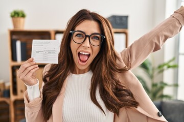 Wall Mural - Young brunette woman holding covid record card celebrating victory with happy smile and winner expression with raised hands