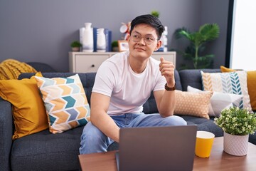 Wall Mural - Young asian man using laptop at home sitting on the sofa smiling looking to the side and staring away thinking.