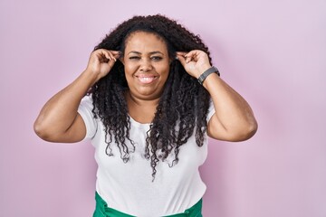 Canvas Print - Plus size hispanic woman standing over pink background smiling pulling ears with fingers, funny gesture. audition problem