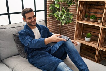 Wall Mural - Young hispanic man watching tv sitting on sofa at home