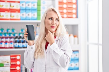 Canvas Print - Caucasian woman working at pharmacy drugstore showing smartphone screen serious face thinking about question with hand on chin, thoughtful about confusing idea