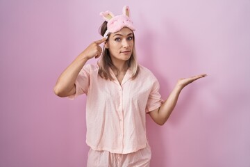 Canvas Print - Blonde caucasian woman wearing sleep mask and pajama confused and annoyed with open palm showing copy space and pointing finger to forehead. think about it.