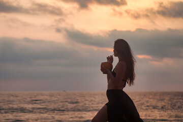 Silhouette elegant woman in black dress drinking coconut on beach at sunset sea background, summer vacation. Lovely lady posing on tropical seacoast. Travel tropic holiday concept. Copy ad text space