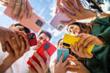 Fototapeta  - Young group of people using mobile phone device standing together in circle outdoors. Millennial friends addicted to social media app, betting or playing video game on platform online.