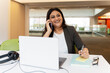© Maria Vitkovska - Confident smiling Indian businesswoman talking on mobile phone, using laptop, working online in modern office. Happy mature secretary answering call, taking notes at workplace. Successful business