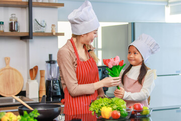 Asian young cheerful female chef housewife mother and little girl daughter wears white tall cook hat and apron standing smiling holding tulip flower bouquet together in kitchen preparing food meal