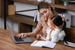 © Bangkok Click Studio - Asian young female housewife mother tutor teacher sitting smiling on table in living room using notebook computer pointing teaching little cute kindergarten preschool girl daughter doing homework