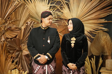 A couple in traditional dress pose for a photo in front of a large wall with a palm tree behind them.