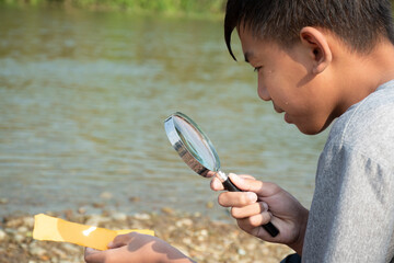 Wall Mural - Asian boy sits holding a magnifying glass and looking at a convex lens to try burning a small piece of paper from the sun's rays near a river in the afternoon during school holidays to do experiment.