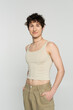 brunette bigender person in tank top standing with hands in pockets of beige pants and smiling at camera isolated on grey.