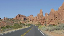 Driving Vehicle On Empty Smooth Curved Road Going Into Distance To Red Mountain Rocks Buttes Landscape In Dried Hot Desert Arches National Park. Highway With Black Asphalt And Orange Road Markings