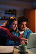 © Jose Calsina - Vertical Young happy caucasian couple browsing on internet using a laptop to search sale at home. Excited husband and cheerful wife smiling and looking for new apartment sitting on desk at livingroom