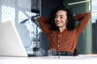 © Liubomir - Happy thinking woman at workplace inside office, business woman with hands behind head completed work well, latin american woman satisfied with achievement results looking at laptop screen.