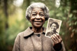 © TRINETTE + CHRIS - Happy senior black woman holding up an old photo of herself when she was young outdoors.
