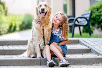 Wall Mural - Preteen girl wearing hat hugging golden retriever dog sitting outdoors in summertime. Pretty kid petting fluffy doggy pet in city