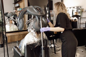 Back view of young woman sitting in front of mirror near hairdresser in beauty salon under hooded dryer machine. Device.