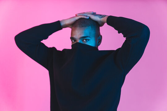 sad black male model with face covered with hoodie collar looking at camera over pink background. mo