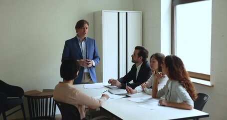 Wall Mural - Team of focused business colleagues, interns listening to middle aged team leader, mentor standing at meeting table, speaking, answering questions, teaching, discussing project plan with group