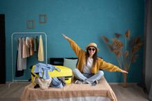 Laughing Young Woman Sitting On Bed With Suitcase At Home, Holding Hands Apart In Flight Ready For Summer Vacation. Traveling Overseas, Plane Flight