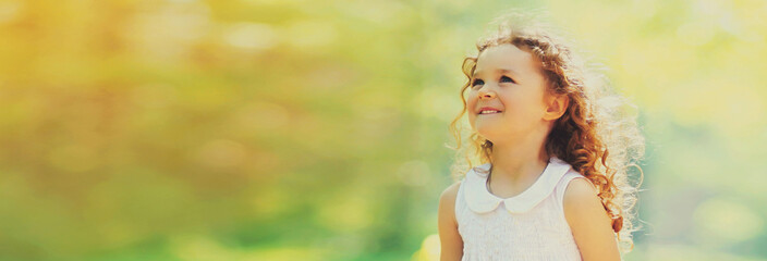 Wall Mural - Portrait of happy cheerful smiling little girl child with curly hair looking up on sunny summer day, blank copy space for advertising text