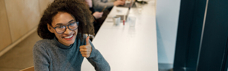 Wall Mural - Smiling woman freelancer working laptop and showing thumb up while sitting in cozy coworking